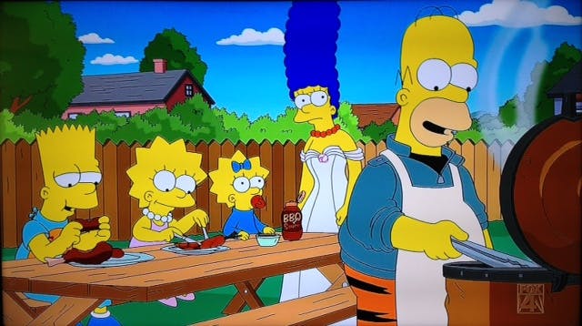 Homer Simpson barbecuing for his family, who sit at a picnic table behind him enjoying the food. 