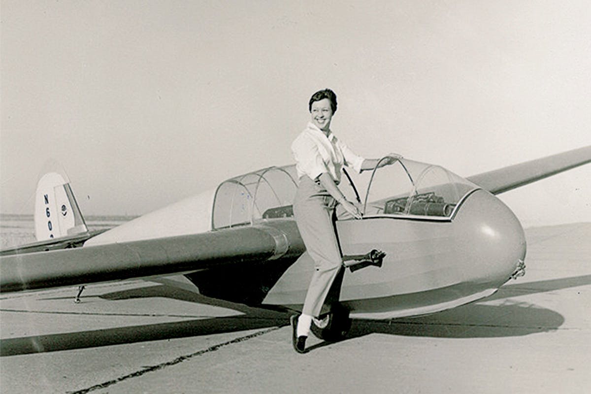 Black and white image of Wally Funk as a young pilot next to a small aircraft. 