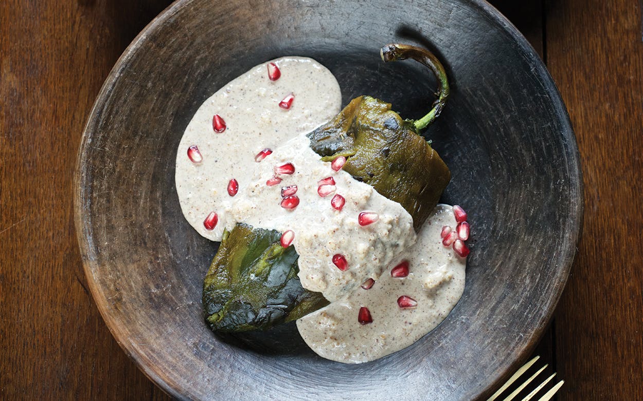 A roasted chile lies in a bowl, covered in a white sauce and pomegranate seeds.