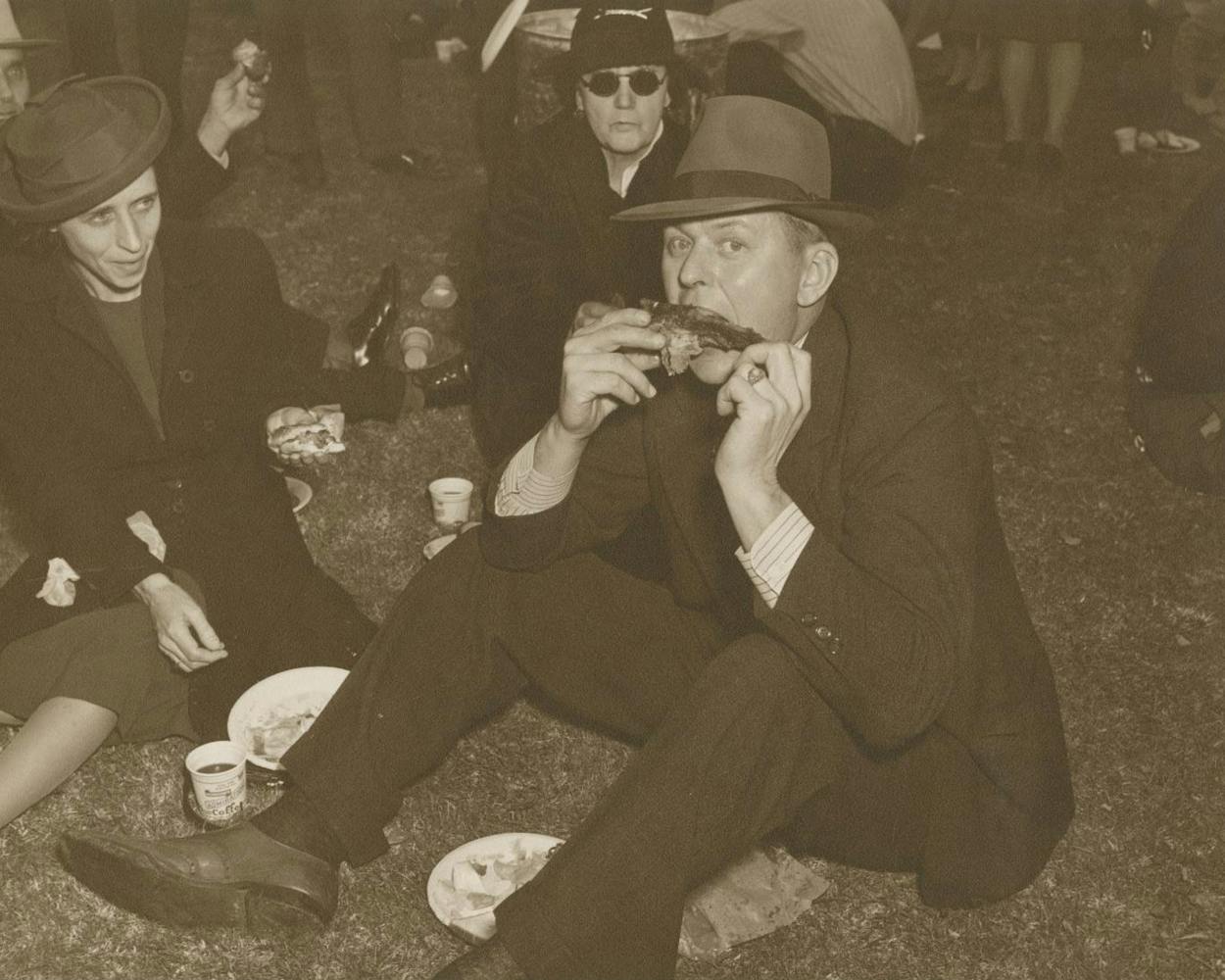 A man sitting on the ground, eating barbecue ribs, in the early 1940s.