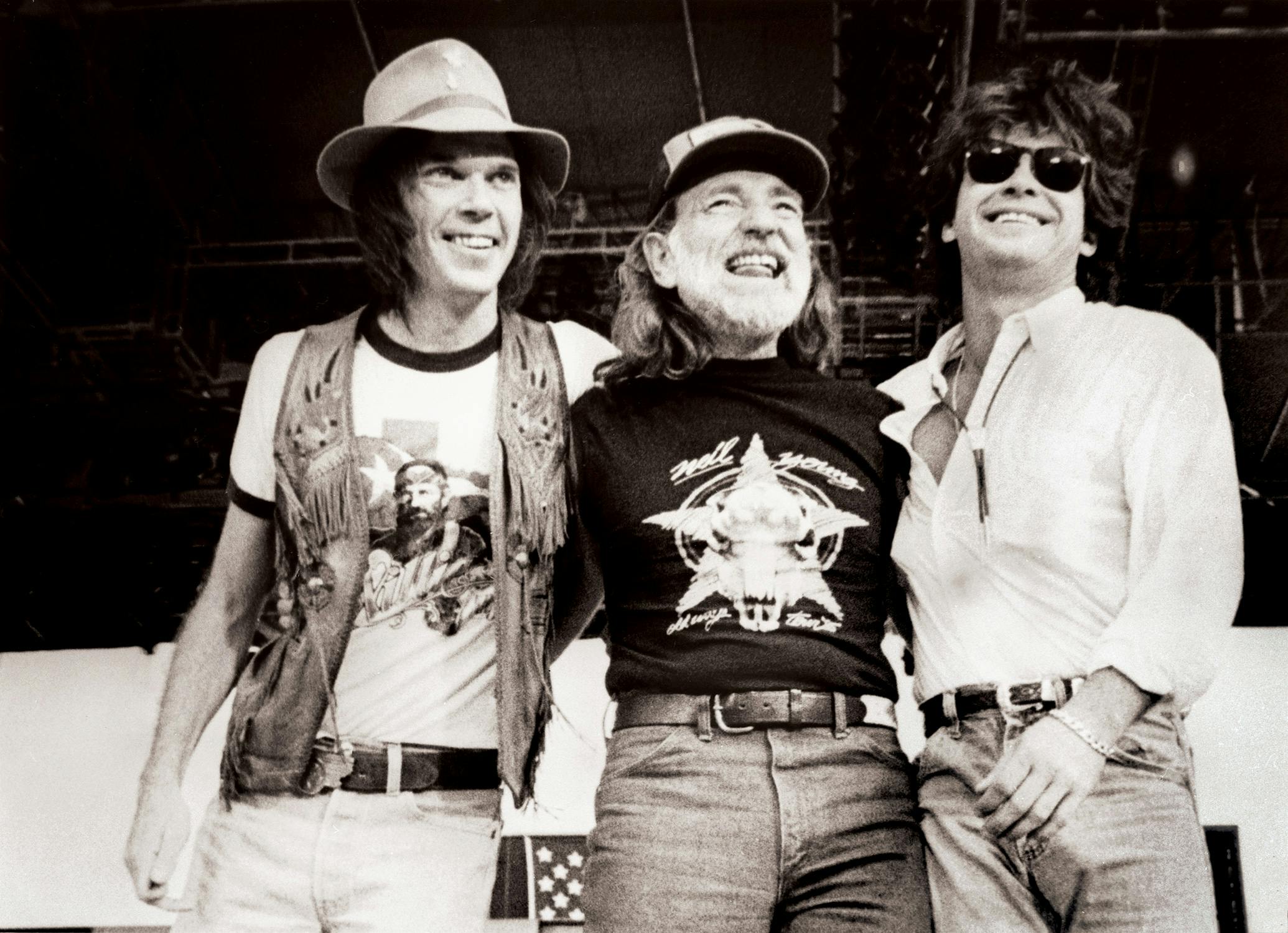 Black and white photograph of Neil Young, Willie, and John Mellencamp smiling on stage. 