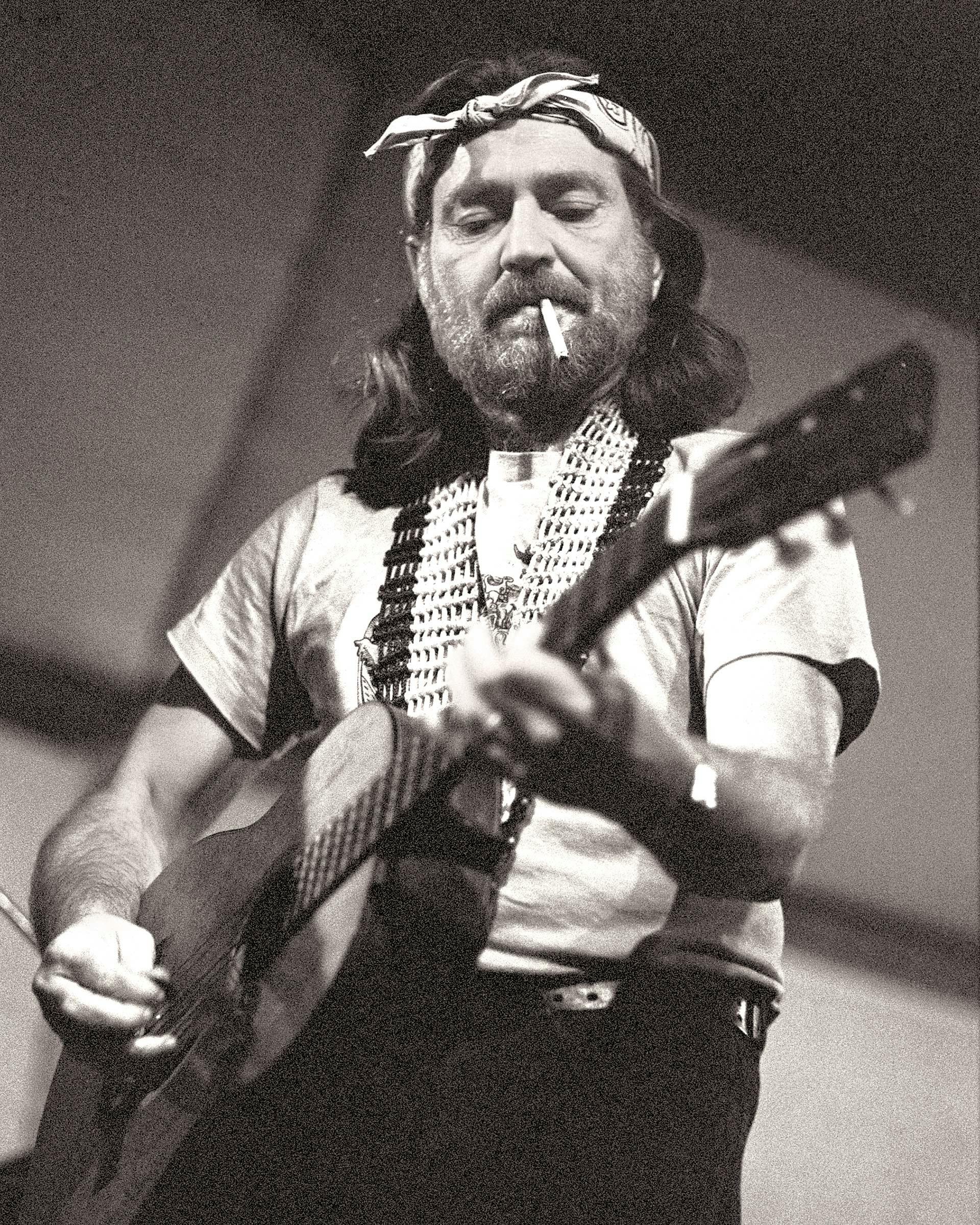 Willie Nelson smoking a cigarette while he strums his guitar. 
