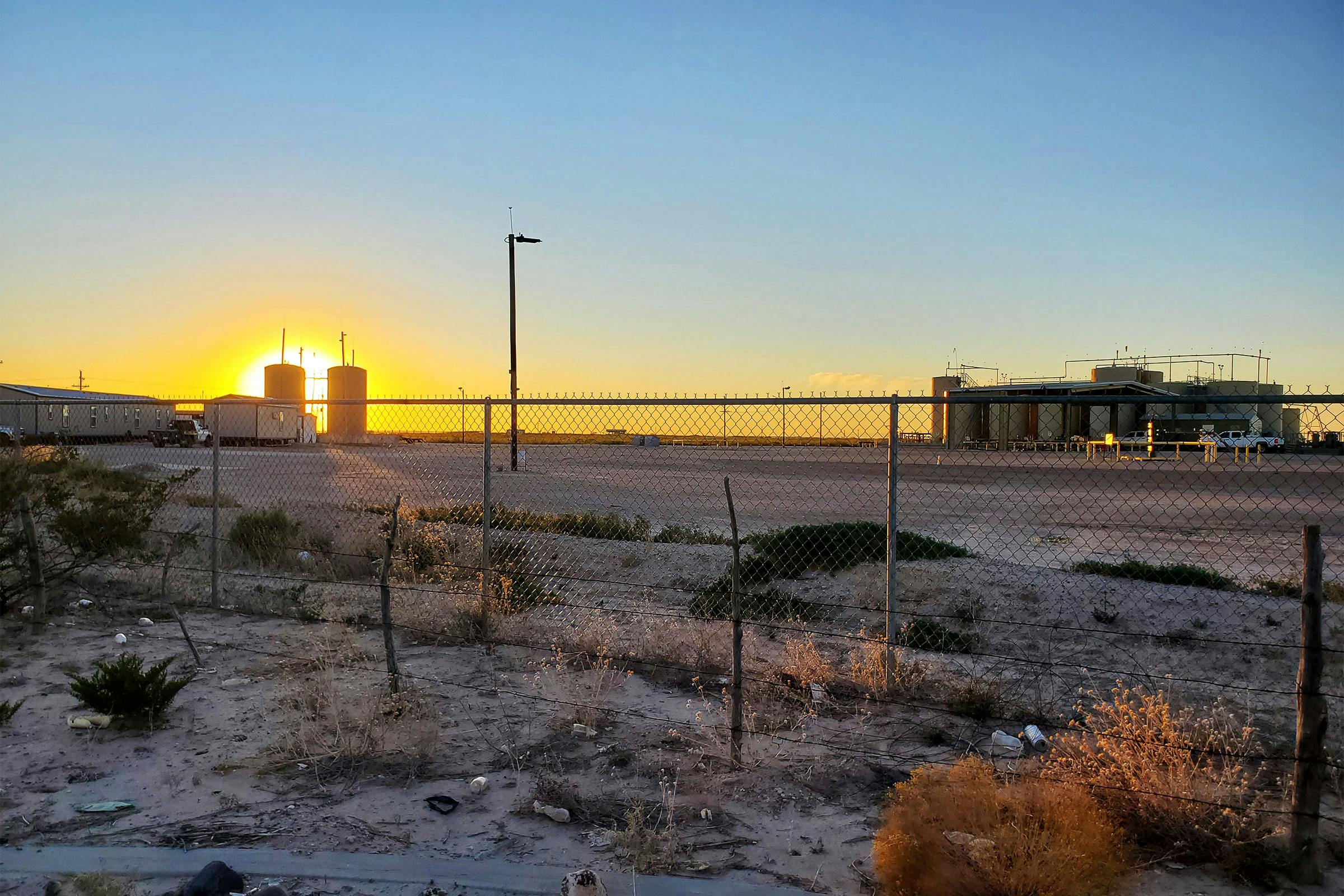 Sunsetting behind tanks and trailers, photographed through a chain link fence. 