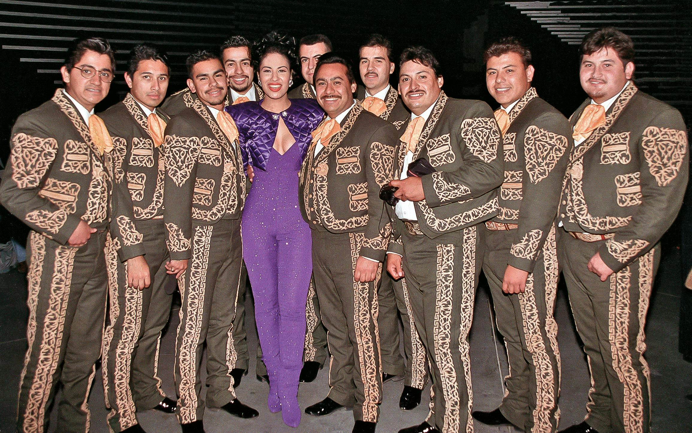 Selena Quintanilla in a purple jumpsuit surrounded by Los Caporales mariachi band. 