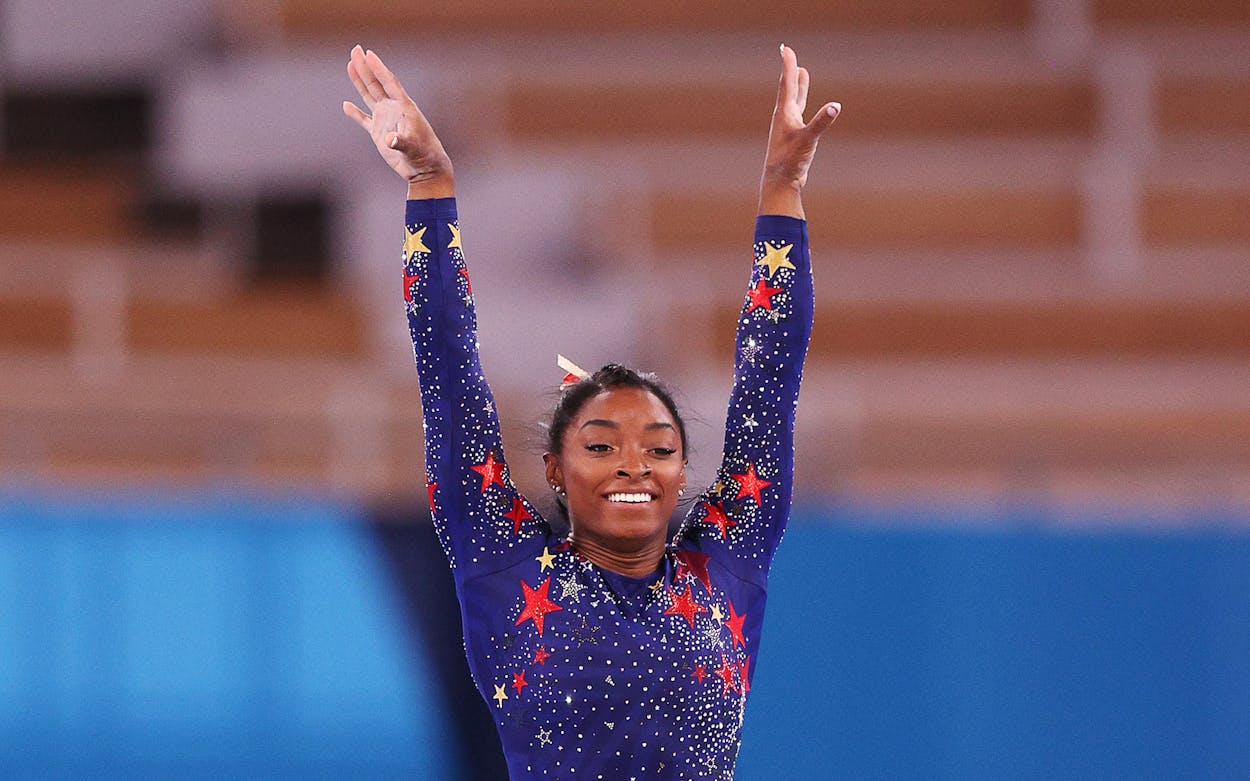 simone biles in a blue, star-covered leotard with her hands stretched above her head.