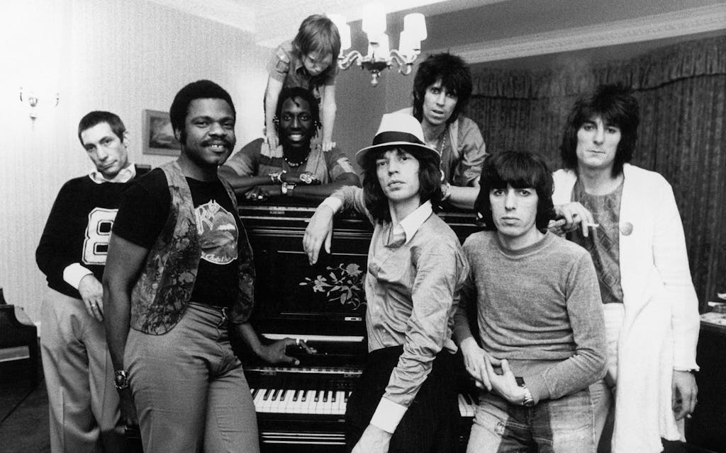 Billy Preston poses with the Rolling Stones in front of a piano in their hotel room. 