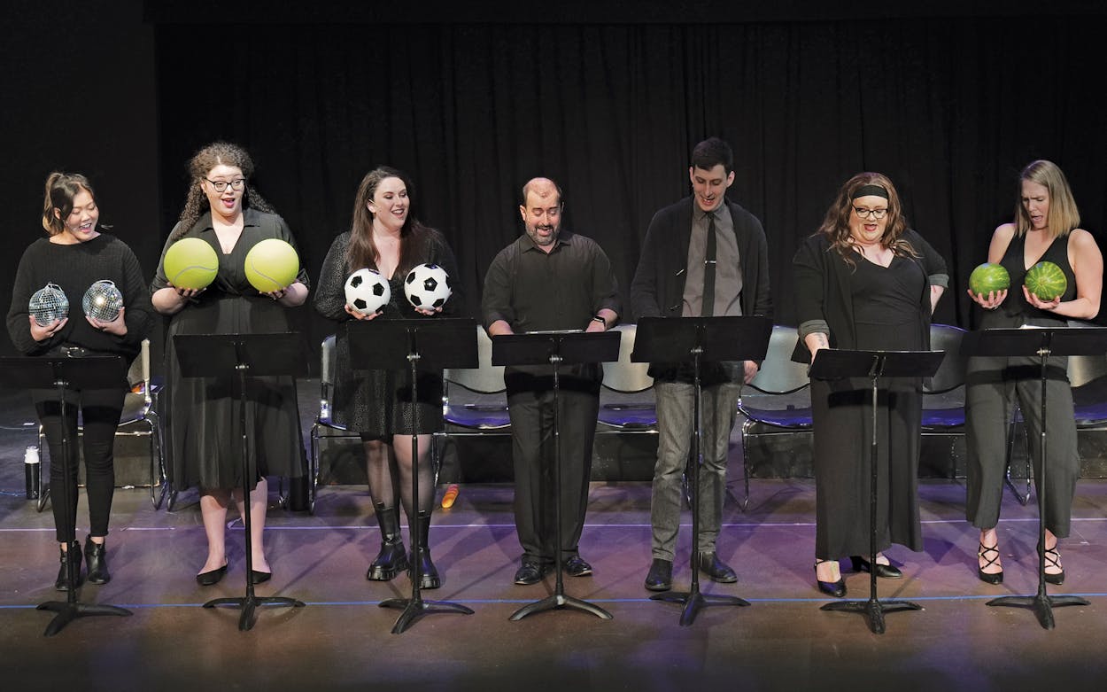 A staged reading of cleaVage: the front story. Seven people are standing on stage holding pairs of round objects up to their chests (disco balls, tennis balls, soccer balls, watermelons, etc.).