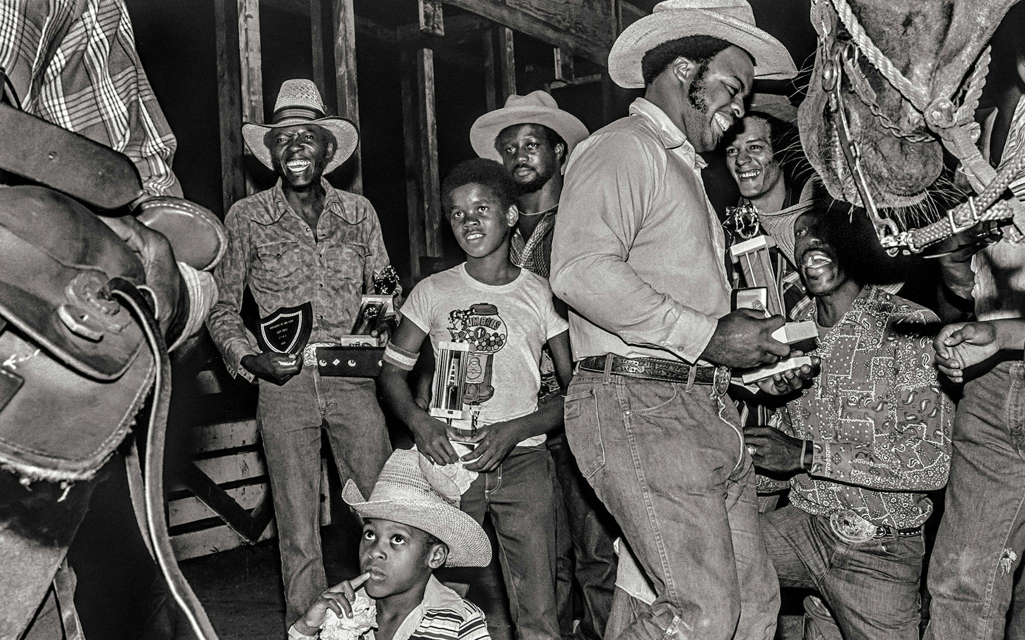 Reginald Page collecting his trophy in 1978 for calf roping while his son, Orlando, also clutching a roping trophy, looks on and rough-stock rider Malloy Scott (second from right) and steer wrestler Jim Richards (kneeling) share a laugh.